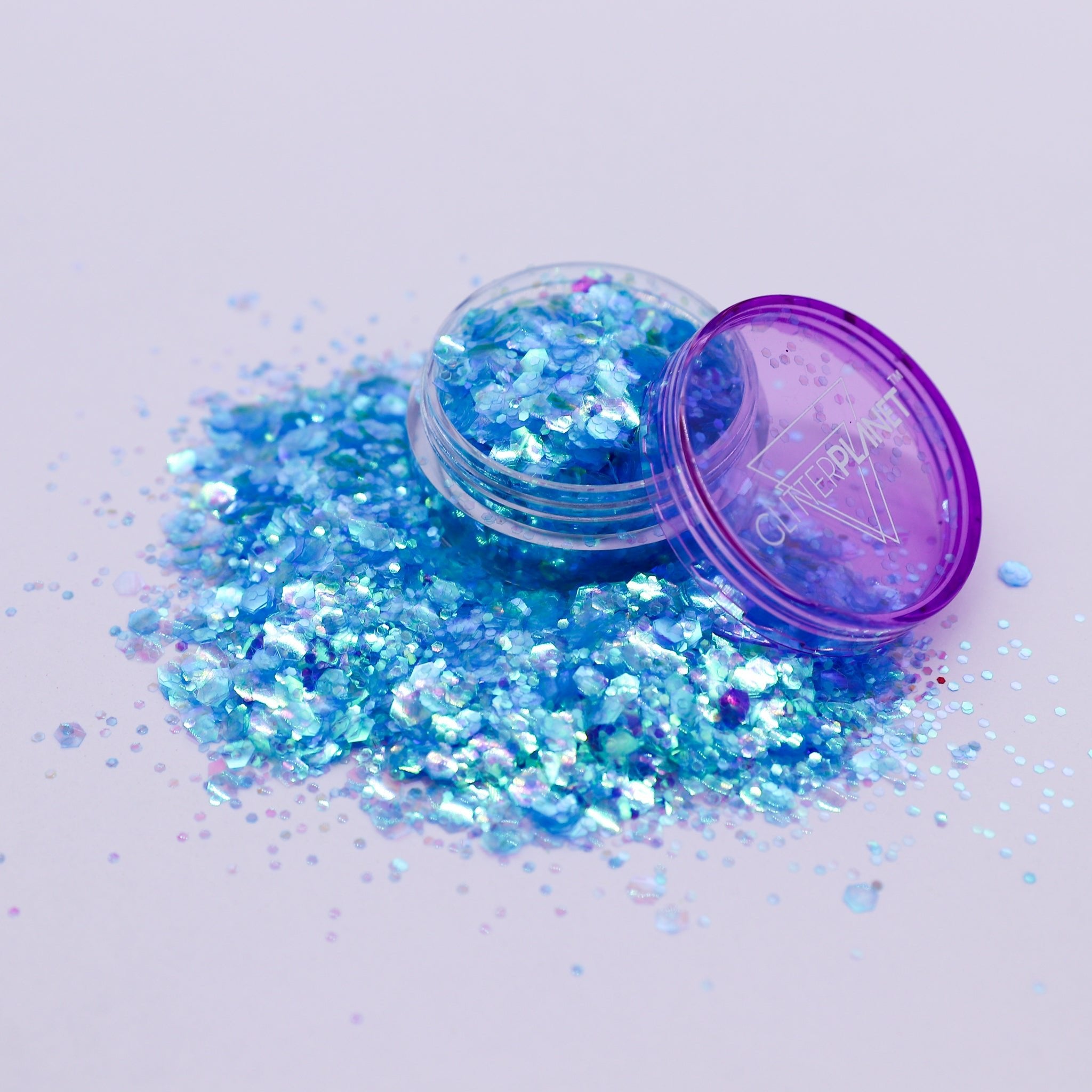 Open container of blue glitter with scattered glitter on a light purple background