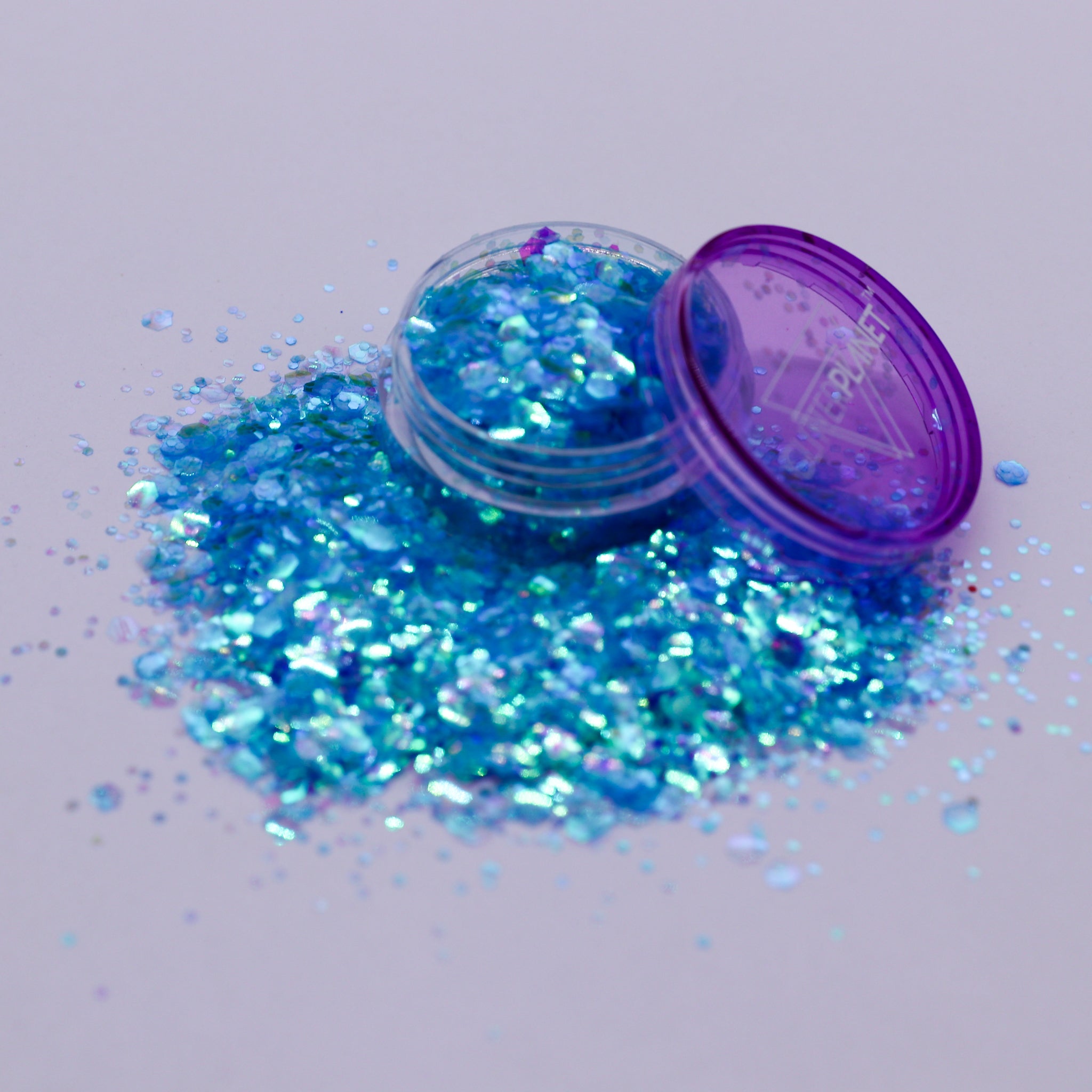 Blue glitter spilling out of a small open container with a purple lid on a light gray background.