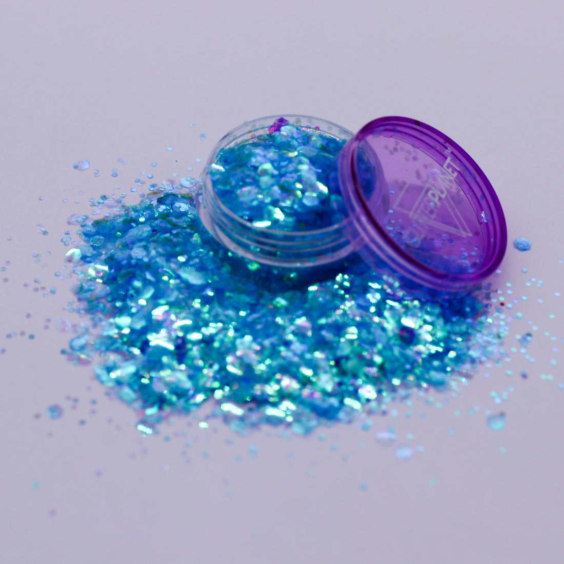 Blue glitter spilling out of a small open container with a purple lid on a light gray background.