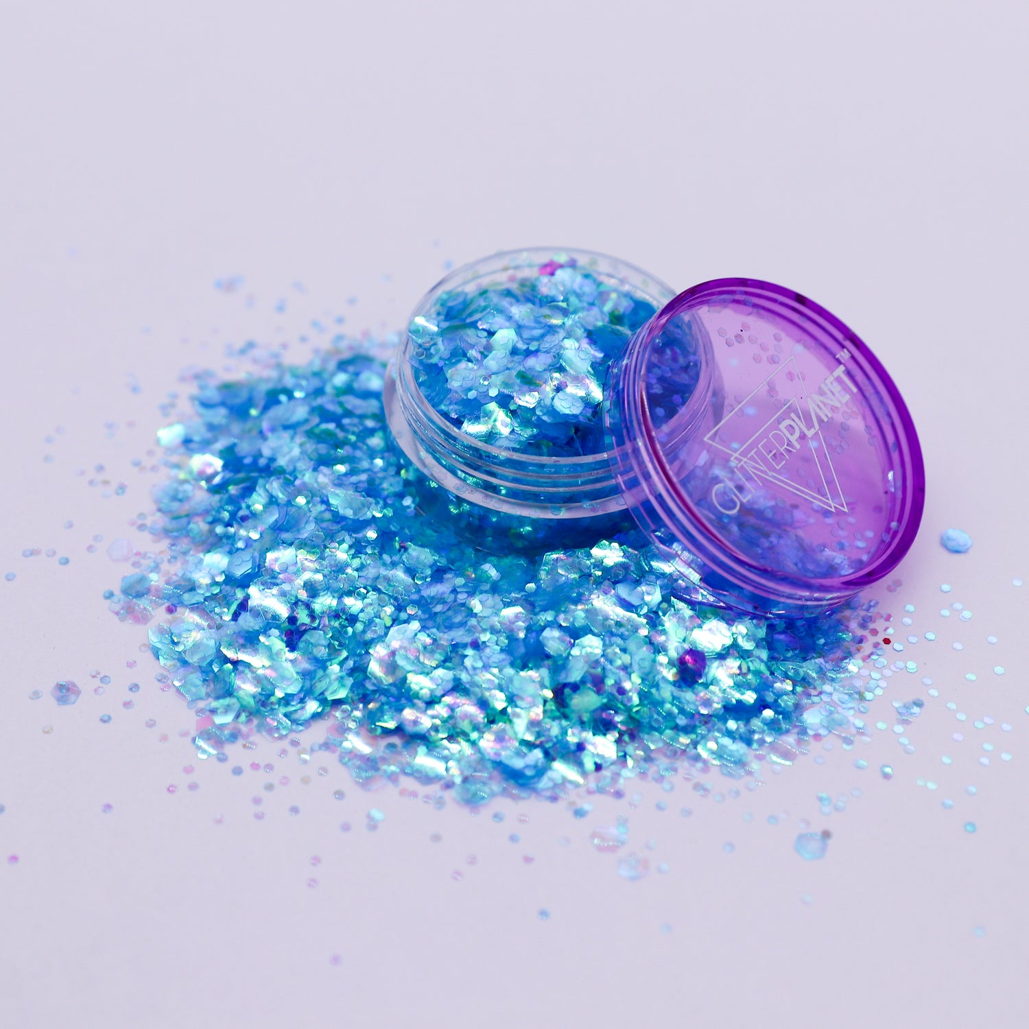 Open container of blue glitter with scattered glitter on a light purple background